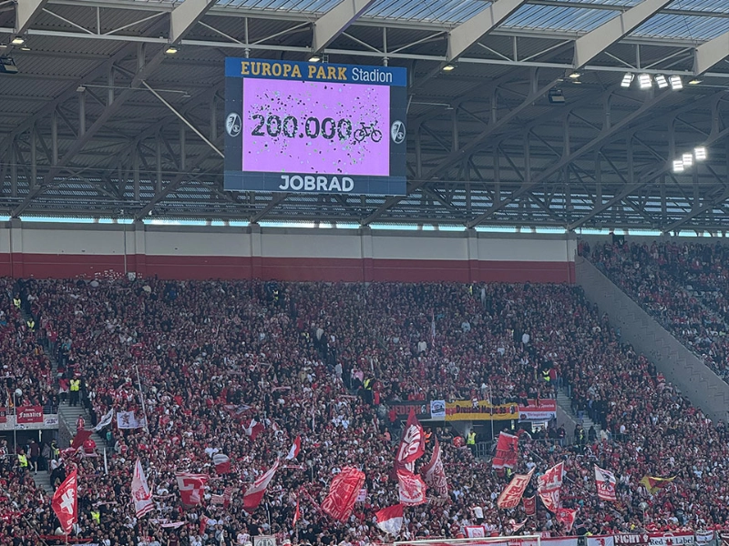 Der Screen im Europa-Park Stadion zeigt an, dass die 200.000 Fahrrad-Fan-Zahl erreicht wurde. Davor sieht man Spieler auf dem Spielfeld. Der Screen im Europa-Park Stadion zeigt an, dass die 200.000 Fahrrad-Fan-Zahl erreicht wurde. Davor sieht man Spieler auf dem Spielfeld.